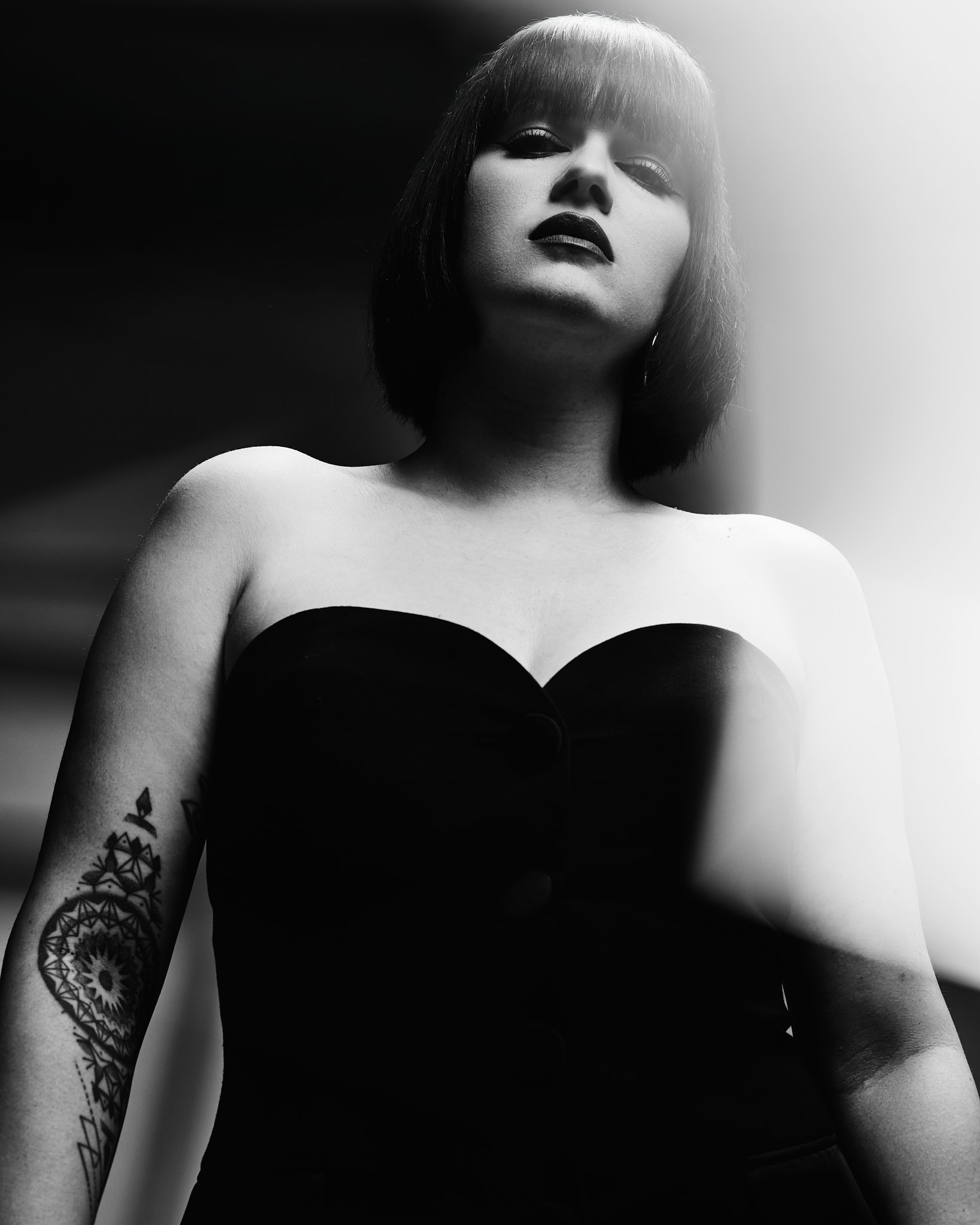 Une femme à la coupe rase et au rouge à lèvres foncé porte un haut noir sans bretelles et se tient debout avec assurance. Son bras tatoué est visible et l'image est en noir et blanc dramatique avec des effets de lumière douce.