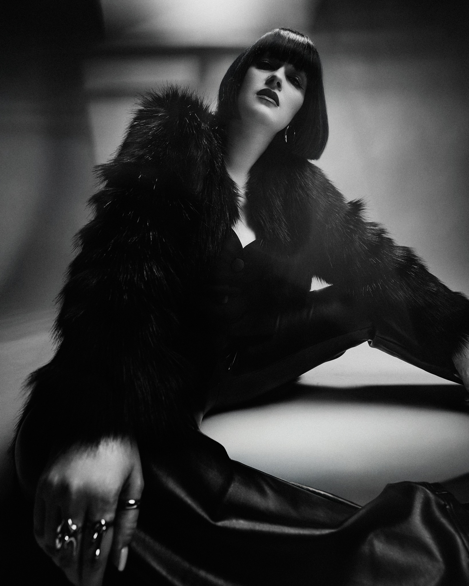 Une femme à la coupe de cheveux lisse est assise sur le sol dans une image dramatique en noir et blanc. Elle porte un manteau en fausse fourrure et un pantalon en cuir, avec une expression confiante et des bagues audacieuses à ses doigts.
