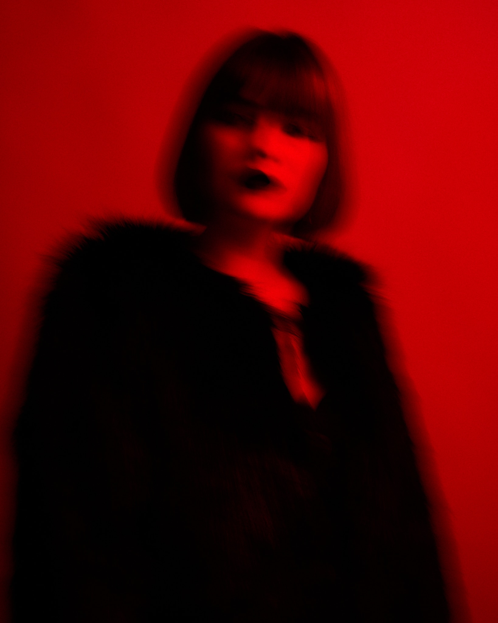 Portrait flou d'une personne coiffée au carré et portant des vêtements sombres, sur un fond rouge vif. L'image a un effet abstrait et lunatique, avec des traits de visage indistincts.