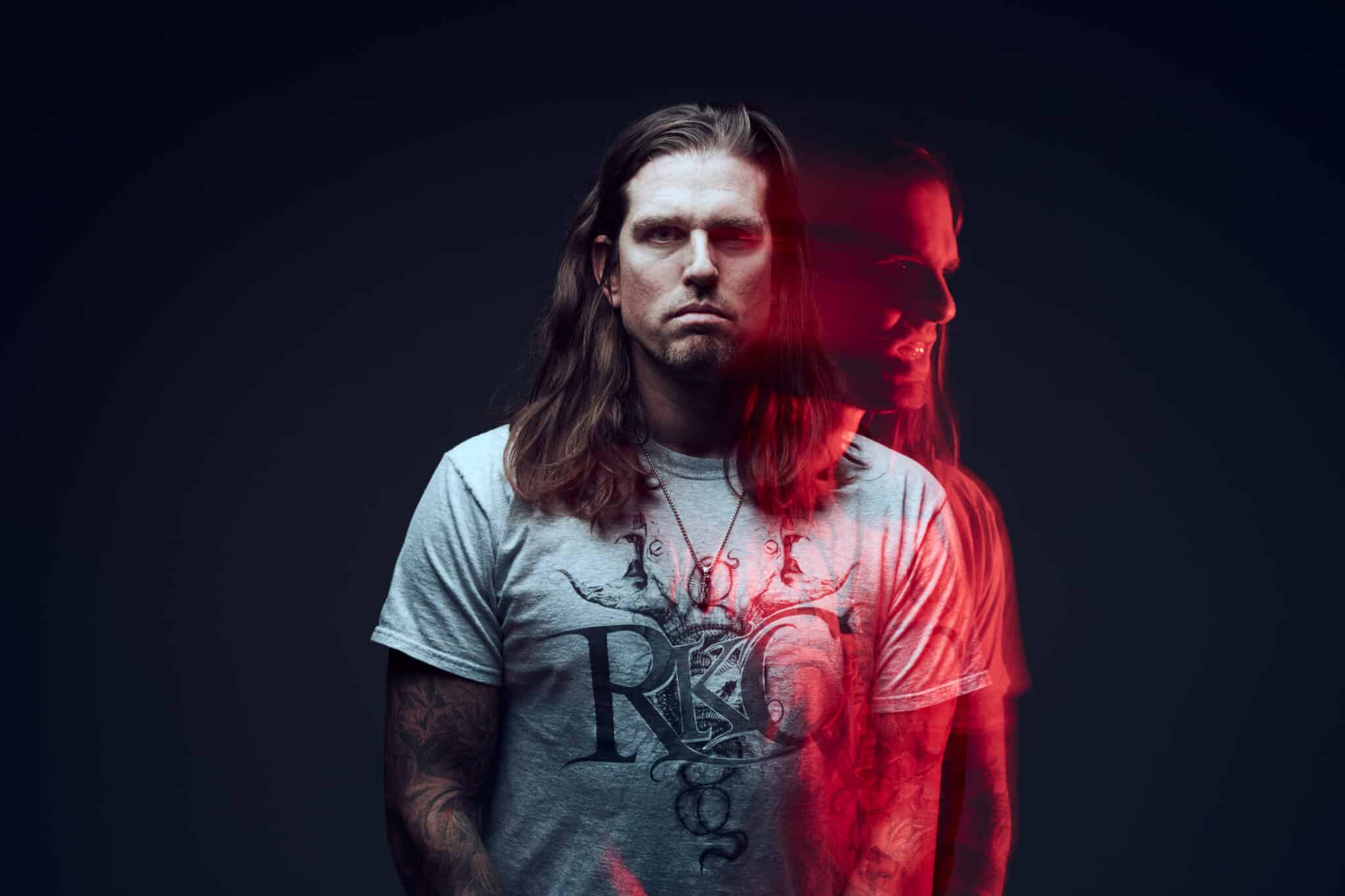 Un homme aux cheveux longs et aux tatouages, vêtu d'un t-shirt clair, se tient debout sur un fond sombre. Une copie fantomatique et teintée de rouge de son visage et de la partie supérieure de son corps apparaît à côté de lui, créant un effet de double exposition.