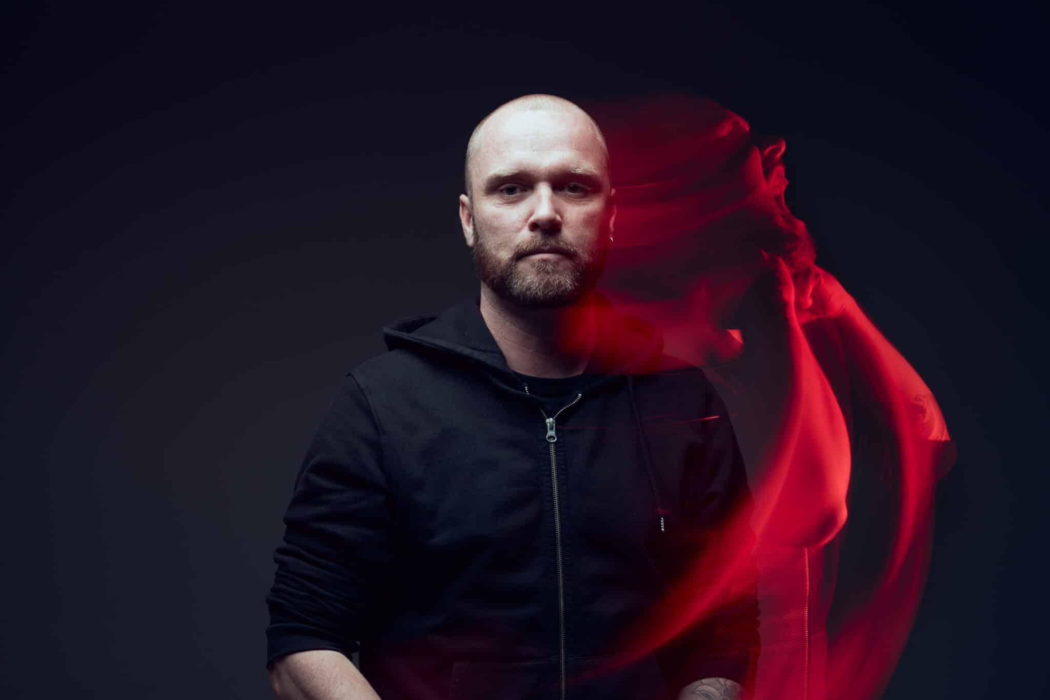Un homme chauve et barbu, vêtu d'un sweat à capuche noir, se tient debout sur un fond sombre. La silhouette fantomatique d'une personne en mouvement, teintée de rouge, apparaît à ses côtés, créant un effet artistique éthéré.
