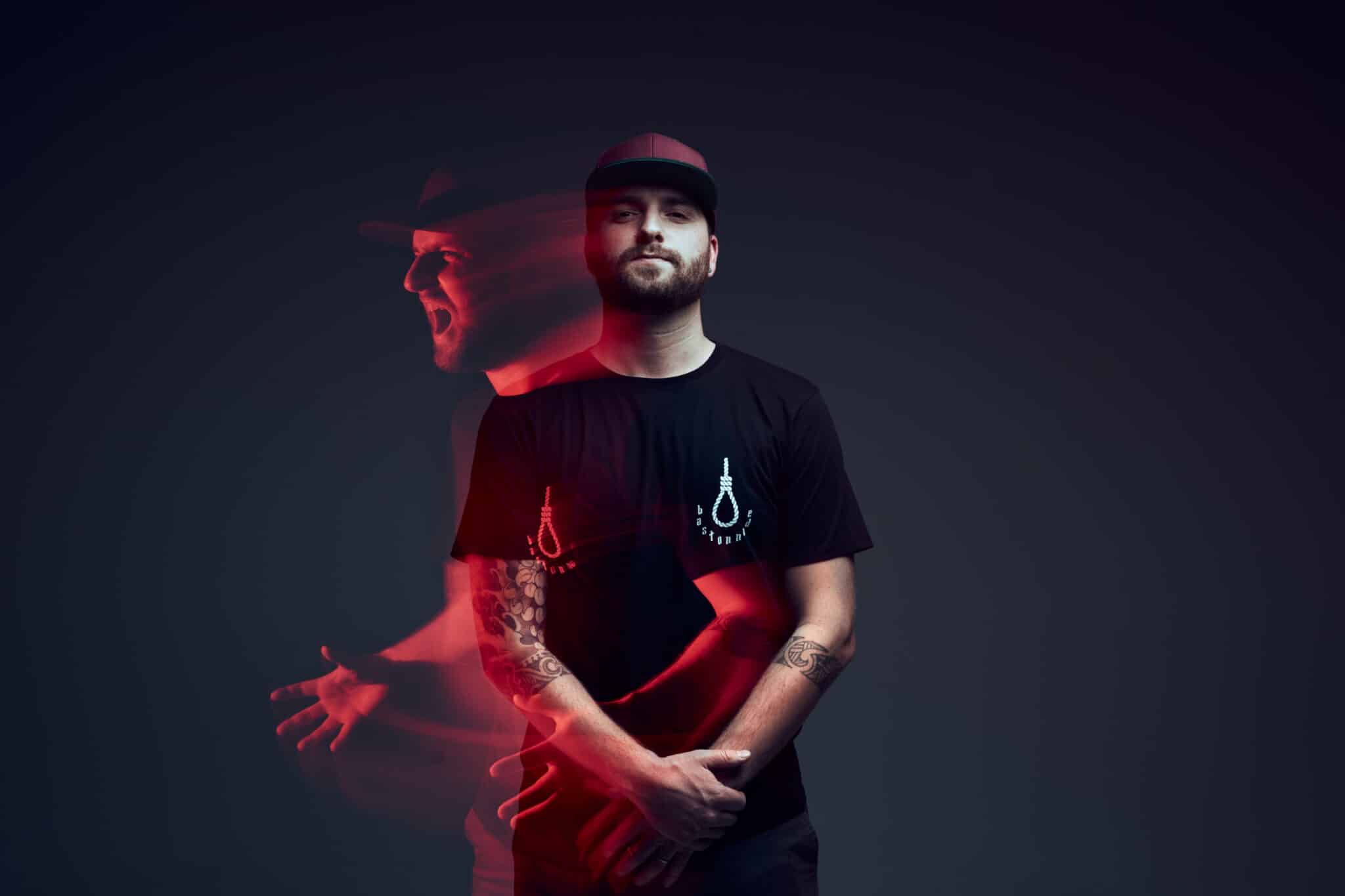 Un homme vêtu d'un t-shirt noir et d'une casquette se tient debout, les bras croisés, tourné vers l'avant. Un flou de bougé teinté de rouge montre une deuxième image de lui à gauche, semblant crier ou hurler, sur un fond dégradé sombre.