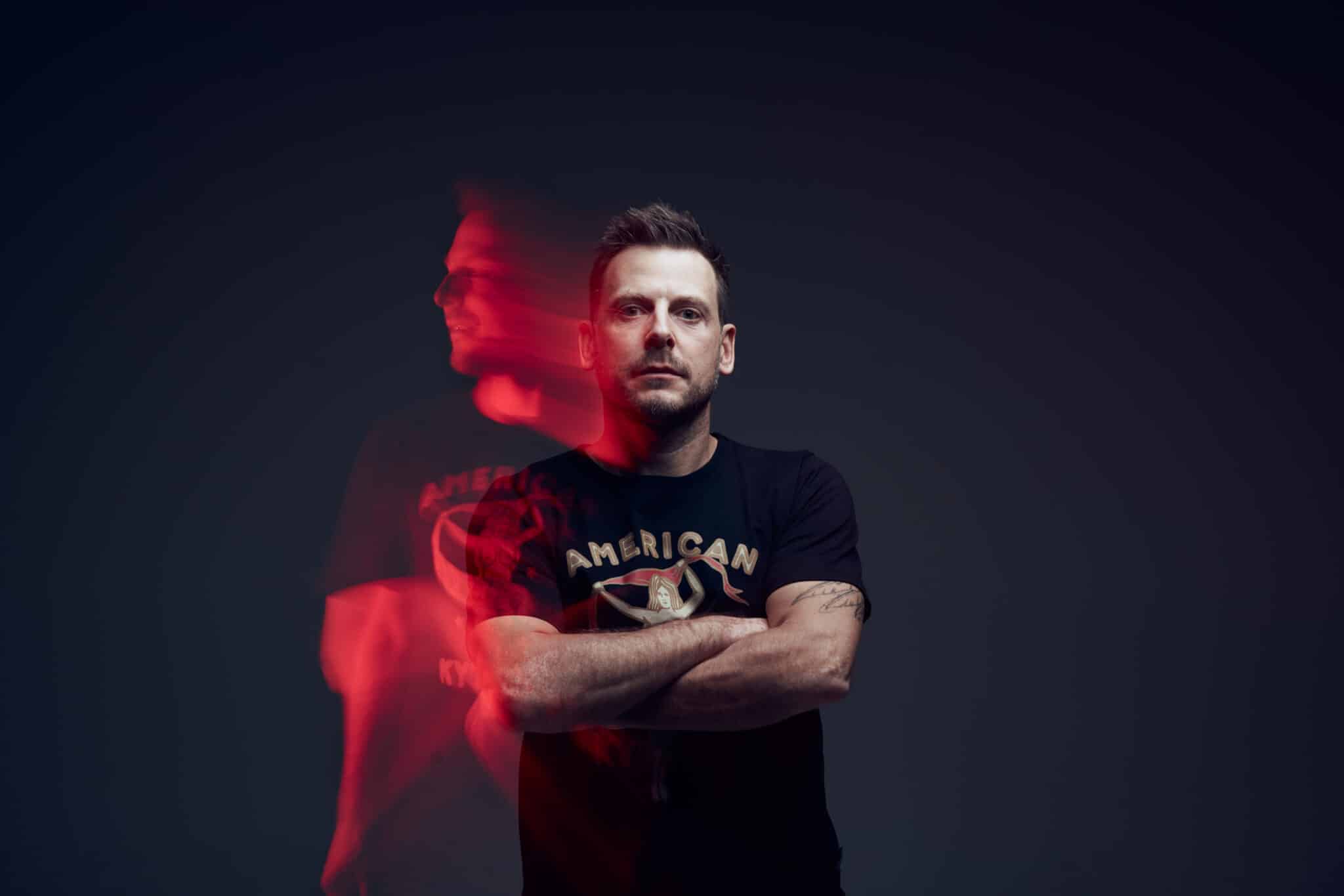 Un homme tatoué se tient debout, les bras croisés, dans une pièce sombre. Il porte un t-shirt américain noir. Un double flou et teinté de rouge de sa silhouette apparaît à sa gauche, créant un effet de mouvement fantomatique.