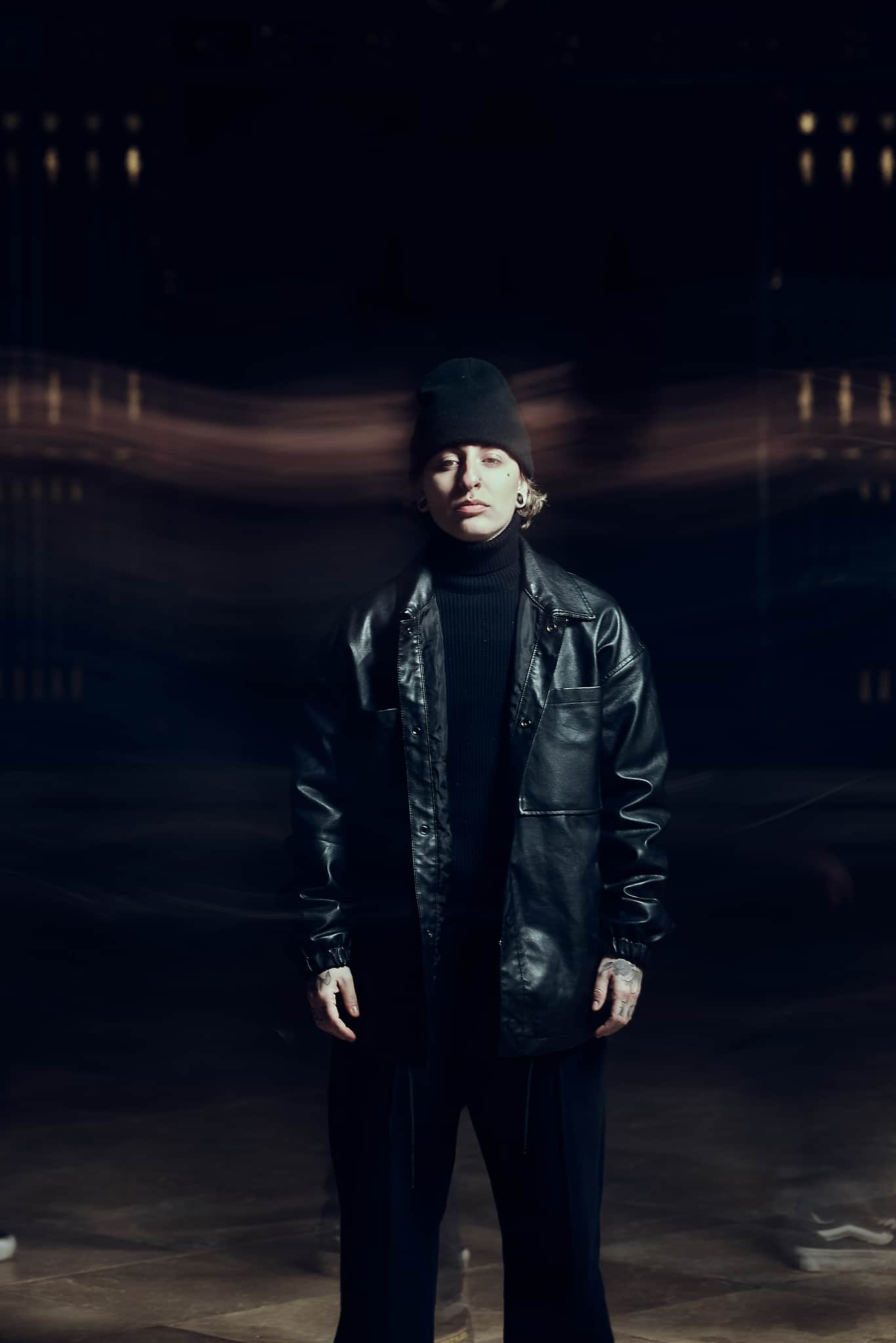 Une personne portant un bonnet noir, un col roulé noir et une veste en cuir noir se tient debout, les yeux fermés, sur un fond sombre et flou avec des raies de lumière.