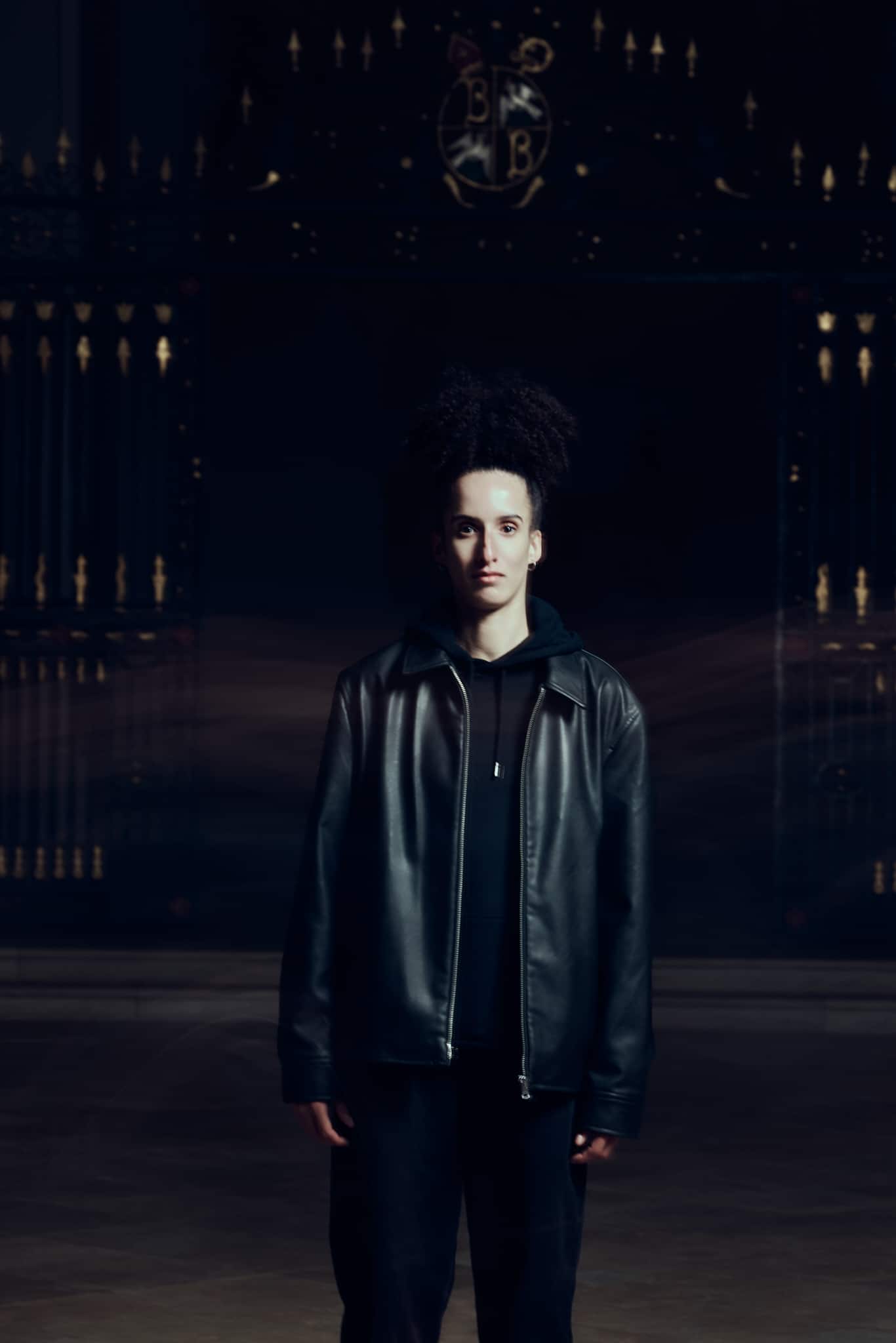 Une personne aux cheveux bouclés se tient face à la caméra, vêtue d'une veste en cuir noir et de vêtements sombres, sur un fond faiblement éclairé avec des détails de portes ornées en noir et or.