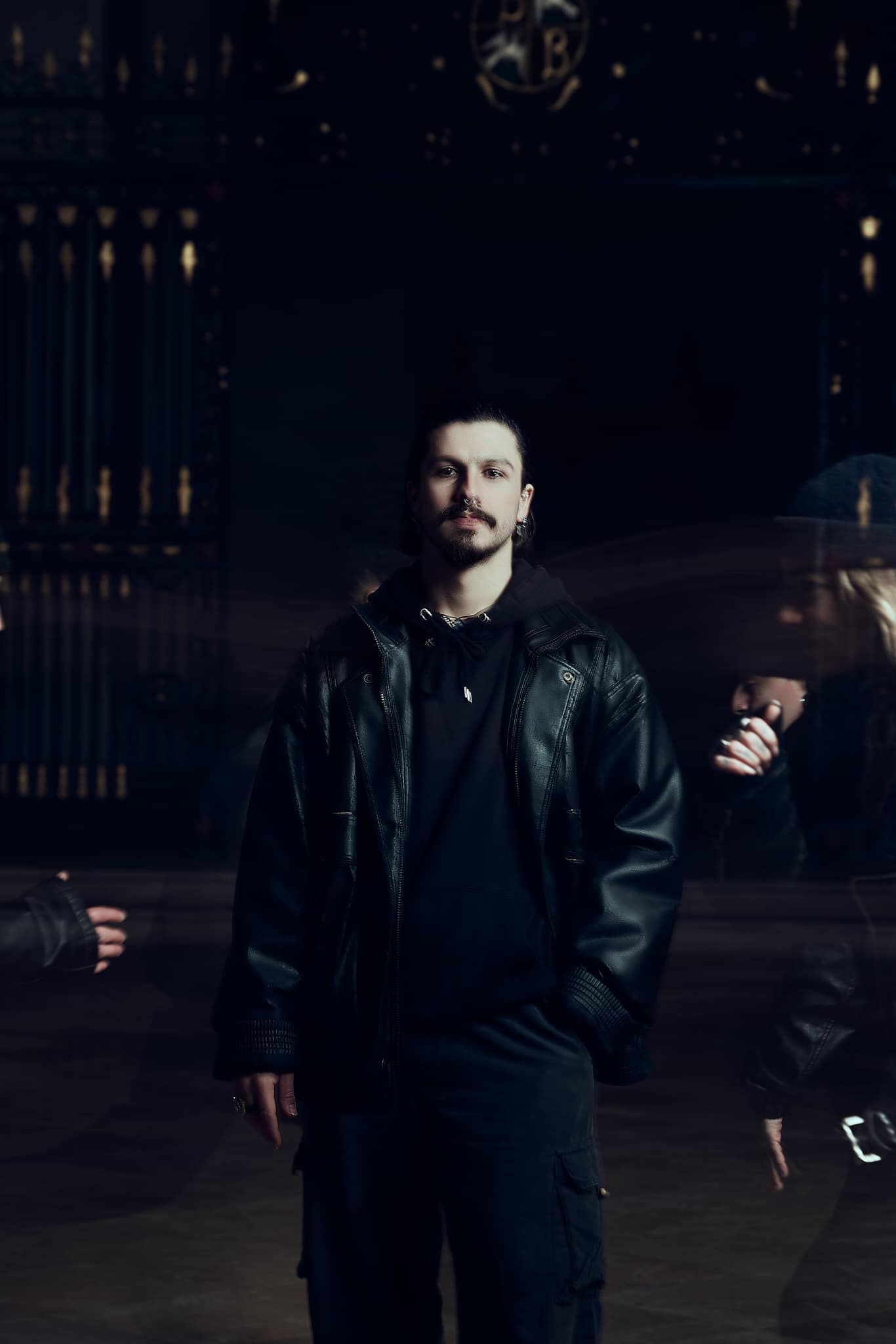 Un homme aux cheveux noirs et à la moustache, vêtu d'un blouson de cuir noir, se tient dans un endroit faiblement éclairé et des silhouettes floues se déplacent autour de lui. L'arrière-plan est orné de ferronneries sombres.