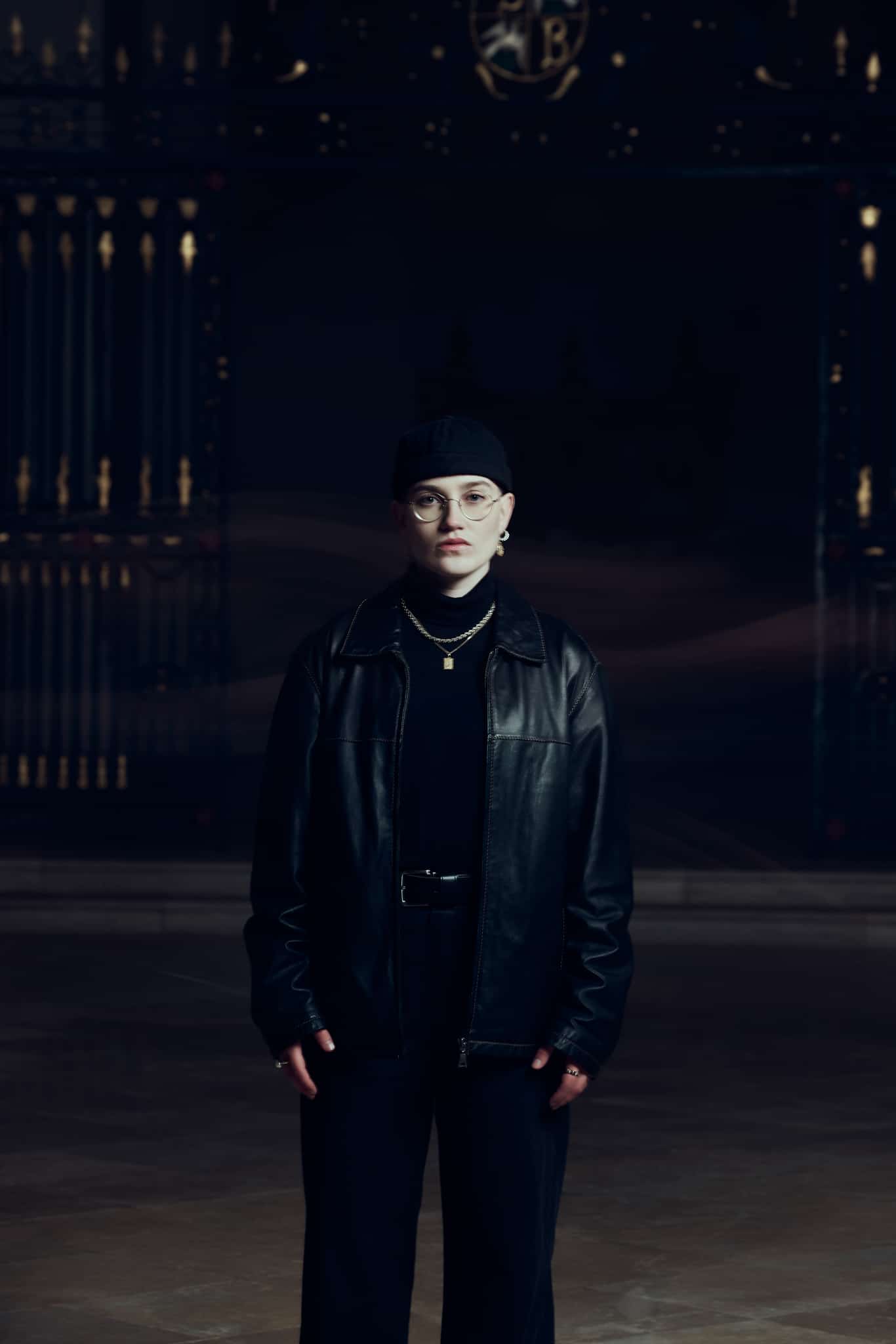Une personne portant des lunettes, un bonnet noir et une veste en cuir noir se tient debout, les mains le long du corps, dans un espace faiblement éclairé, avec des grilles métalliques ornées en arrière-plan.