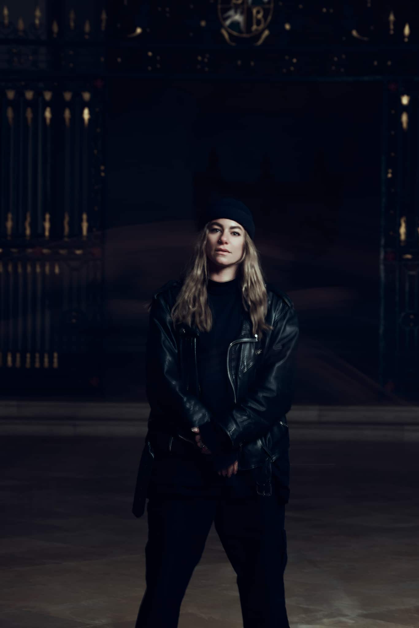 Une personne aux cheveux longs portant un bonnet noir, une veste en cuir noir et un pantalon noir se tient debout, les mains jointes devant elle, sur un fond sombre et orné.