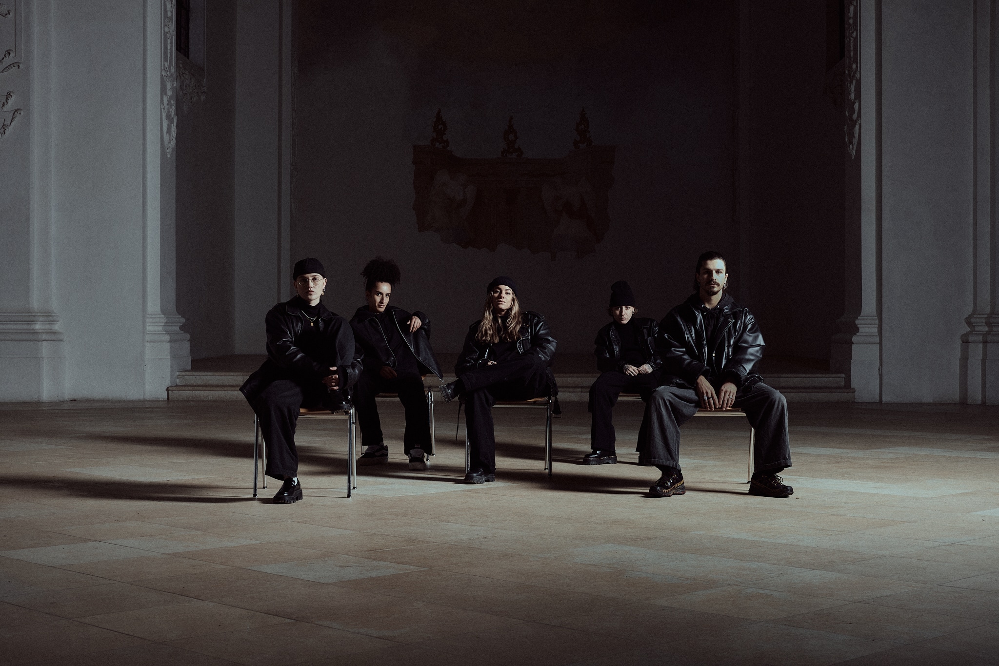 Five individuals dressed in dark clothing are seated on chairs in a spacious, dimly lit room with white, ornate walls and a mural in the background. The group's expressions are serious, and the atmosphere is somber.
