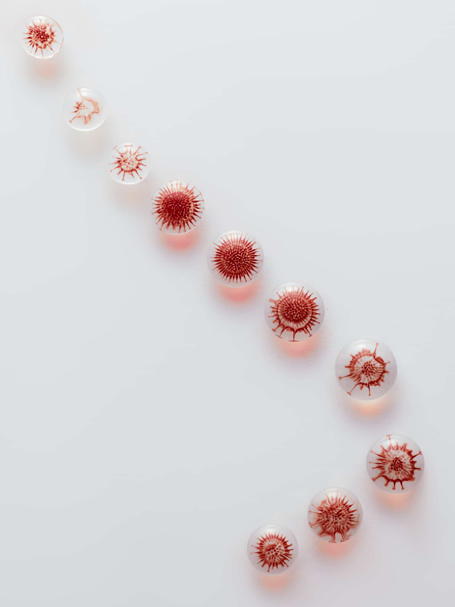 Dix billes en verre transparent, à l'intérieur desquelles se trouvent des motifs rouges ressemblant à des virus, sont disposées en diagonale sur un fond blanc, créant ainsi un motif visuellement frappant et ordonné.