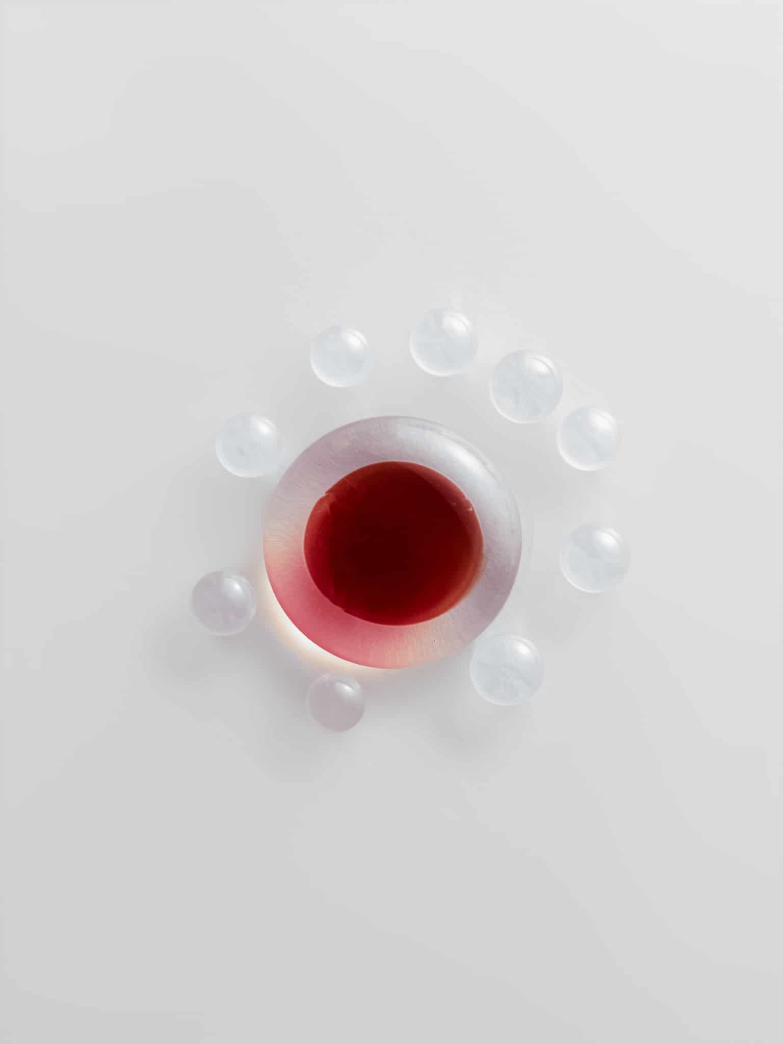 Macro. Une coupelle ronde en verre avec un liquide rouge foncé au centre, entouré d'un anneau de perles translucides et sphériques en forme de gel sur un fond blanc.