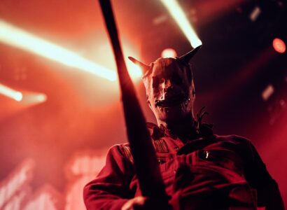 Une personne portant un masque métallique à cornes et une combinaison rouge se tient sur scène sous un éclairage dramatique, à propos d'un spectacle électrisant dont les faisceaux lumineux illuminent l'arrière-plan.
