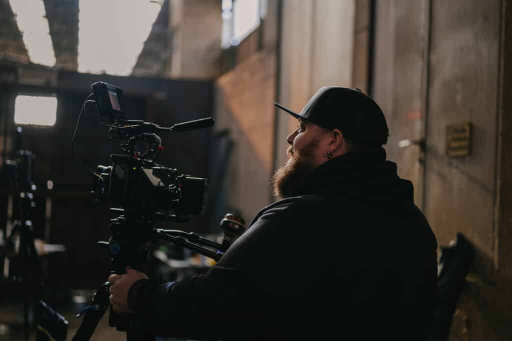A bearded man in a hoodie and black cap operates a professional video camera indoors, in an industrial setting where warm light streams through the windows—a scene to propose for a creative narrative.