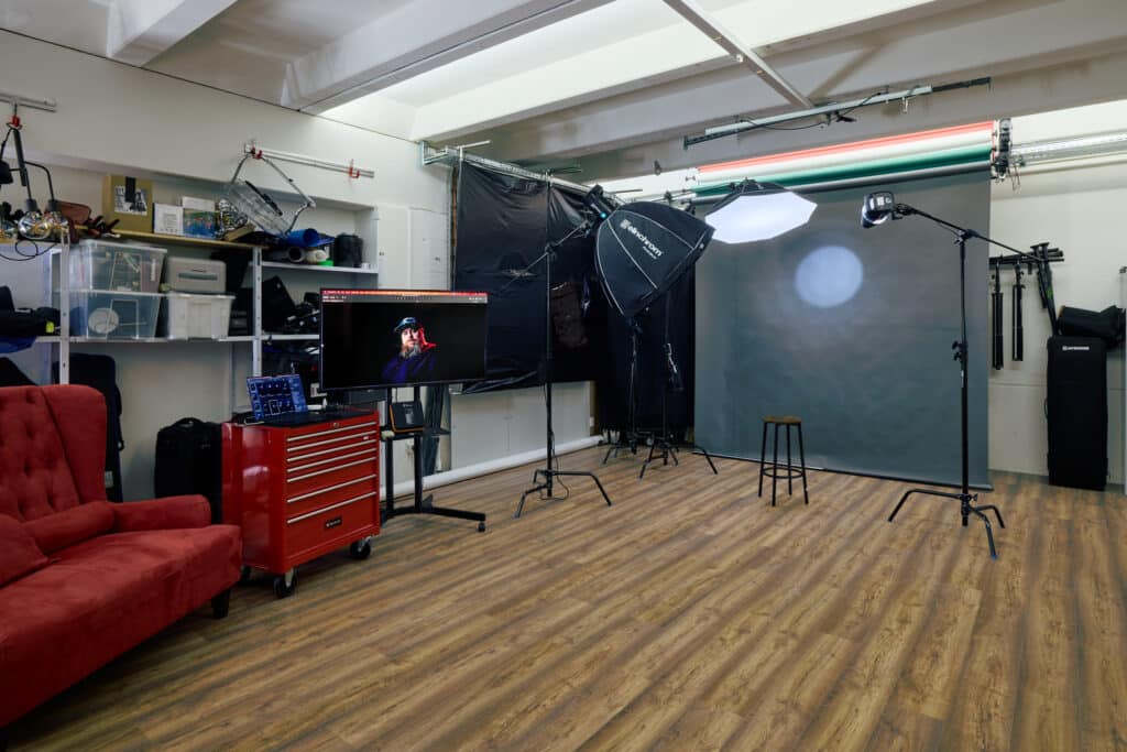 A photographer's about studio featuring lighting equipment, a gray backdrop, a stool, a red toolbox, and a comfortable red sofa. Shelves contain various equipment, while a computer screen on the left displays a portrait photograph.