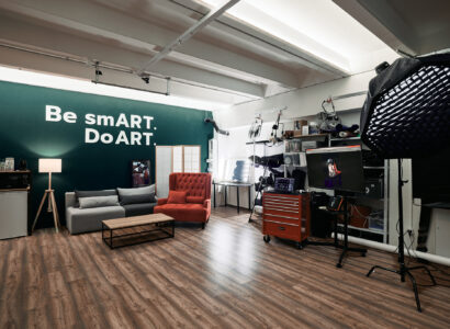 Un studio photo moderne avec un sol en bois, un mur d'accent vert portant l'inscription "Be smART. Do ART.", un canapé gris, un fauteuil rouge, des lampes de studio, du matériel photographique et un ordinateur affichant un portrait.