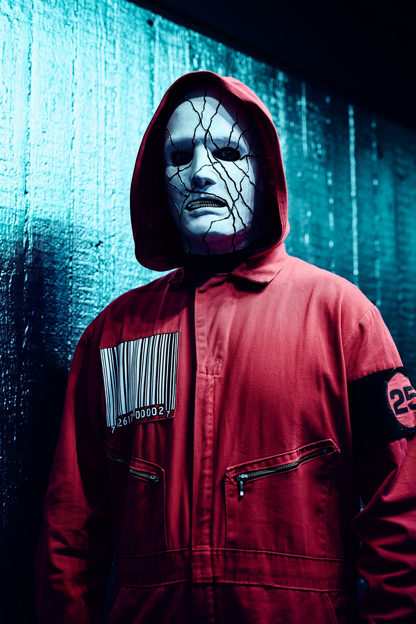Une personne porte un masque blanc craquelé rappelant Slipknot et une combinaison à capuche rouge avec un grand code-barres sur la poitrine et 25 sur la manche, debout contre un mur bleu texturé.