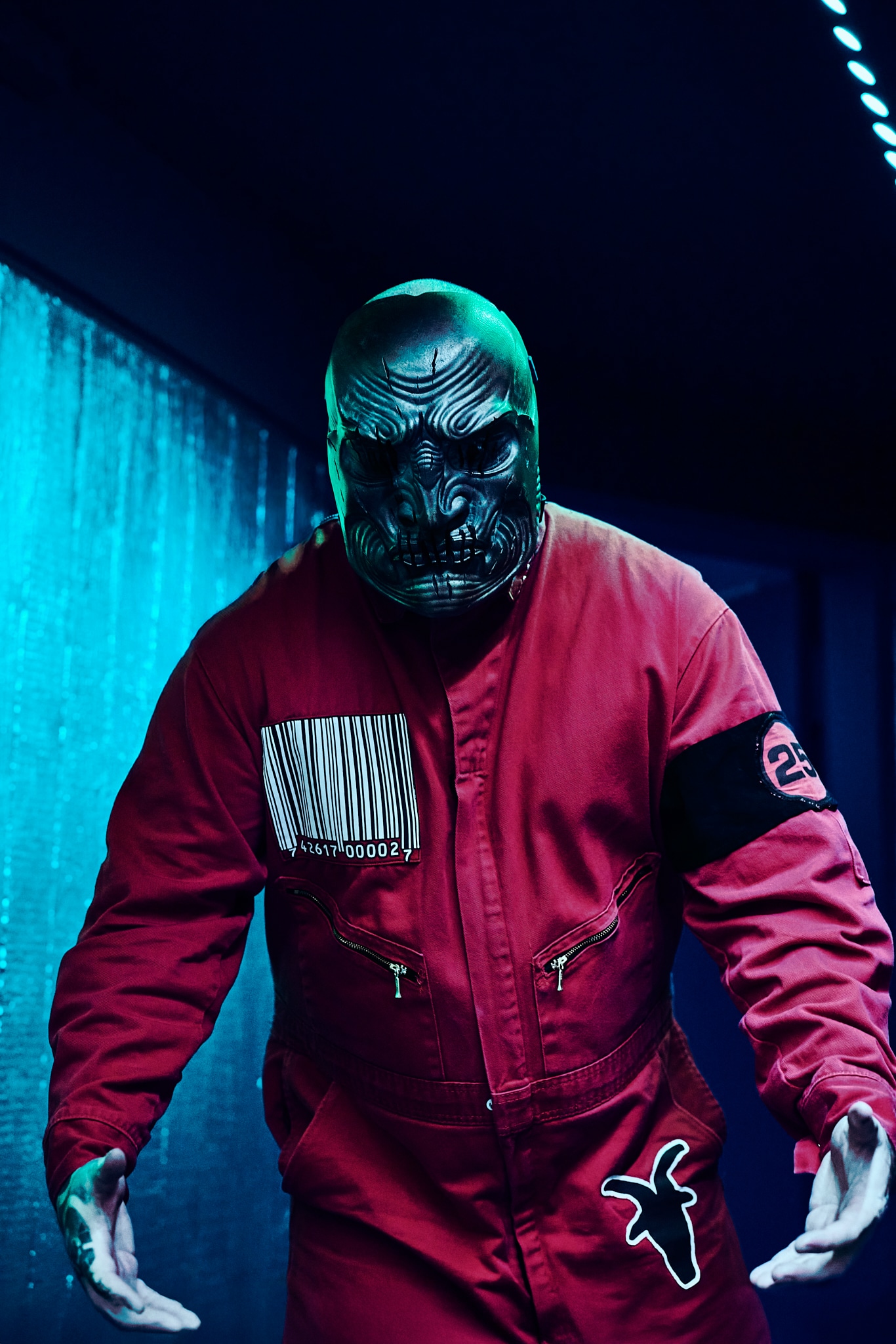 Une personne portant un masque noir détaillé et menaçant rappelant Slipknot et une combinaison rouge avec un code-barres, le chiffre 23 et un symbole blanc se tient sous un éclairage bleu, les bras légèrement tendus.