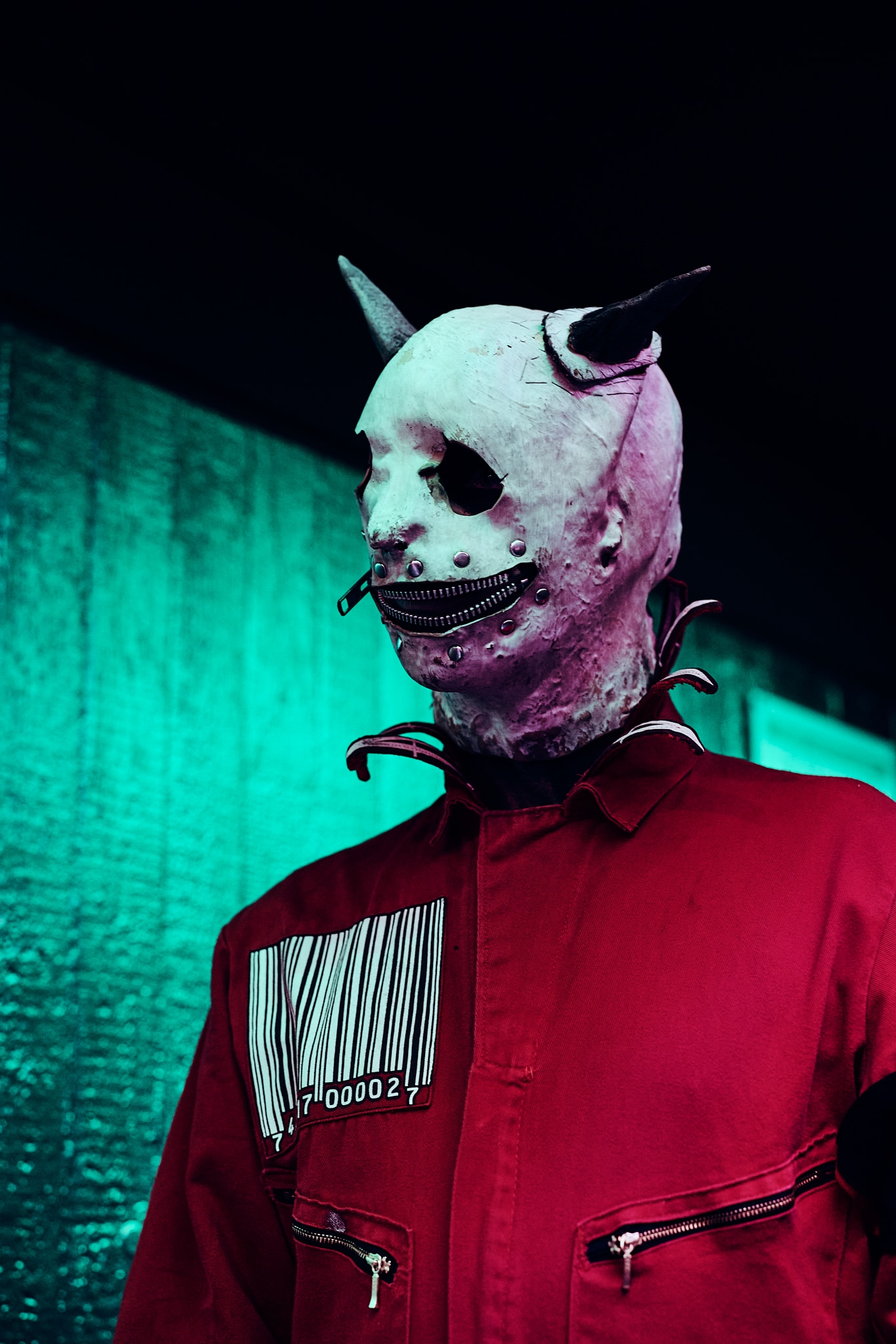 Une personne portant une veste rouge avec un code-barres et un masque à cornes effrayant, inspiré de Slipknot et couvert de clous métalliques, se tient sur un fond éclairé en vert.