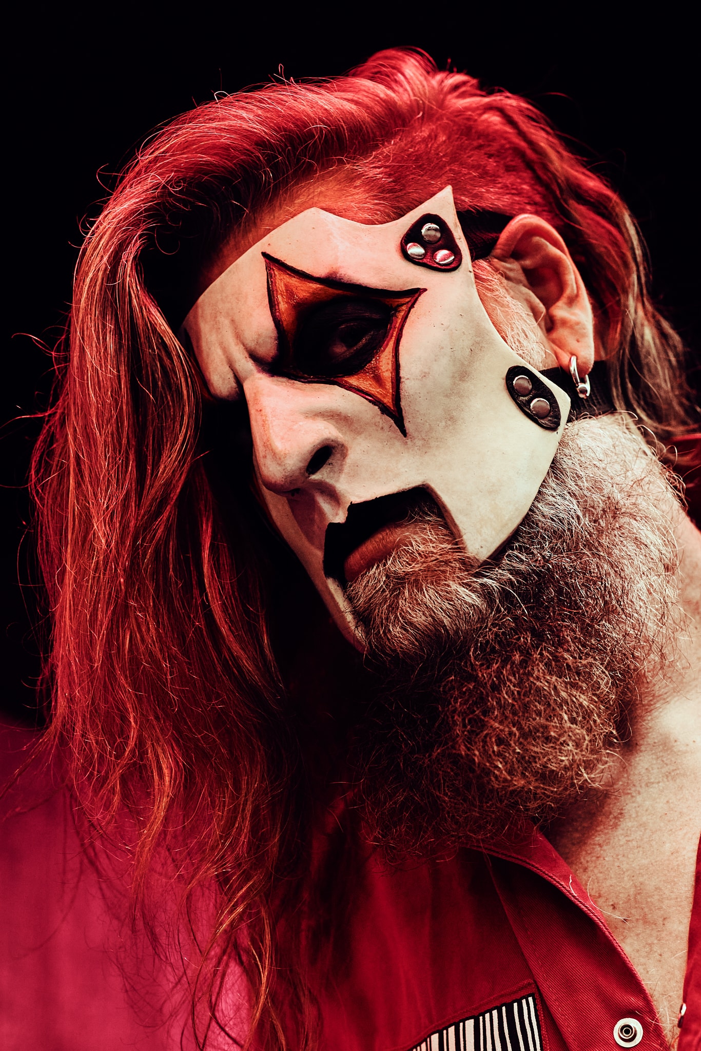 Une personne aux cheveux longs et à la barbe épaisse porte un maquillage de clown théâtral rappelant celui de Slipknot, avec des étoiles sombres autour d'un œil et des ombres dramatiques. Vêtus de rouge et baignés dans un éclairage rouge, ils dégagent une atmosphère intense et morose.