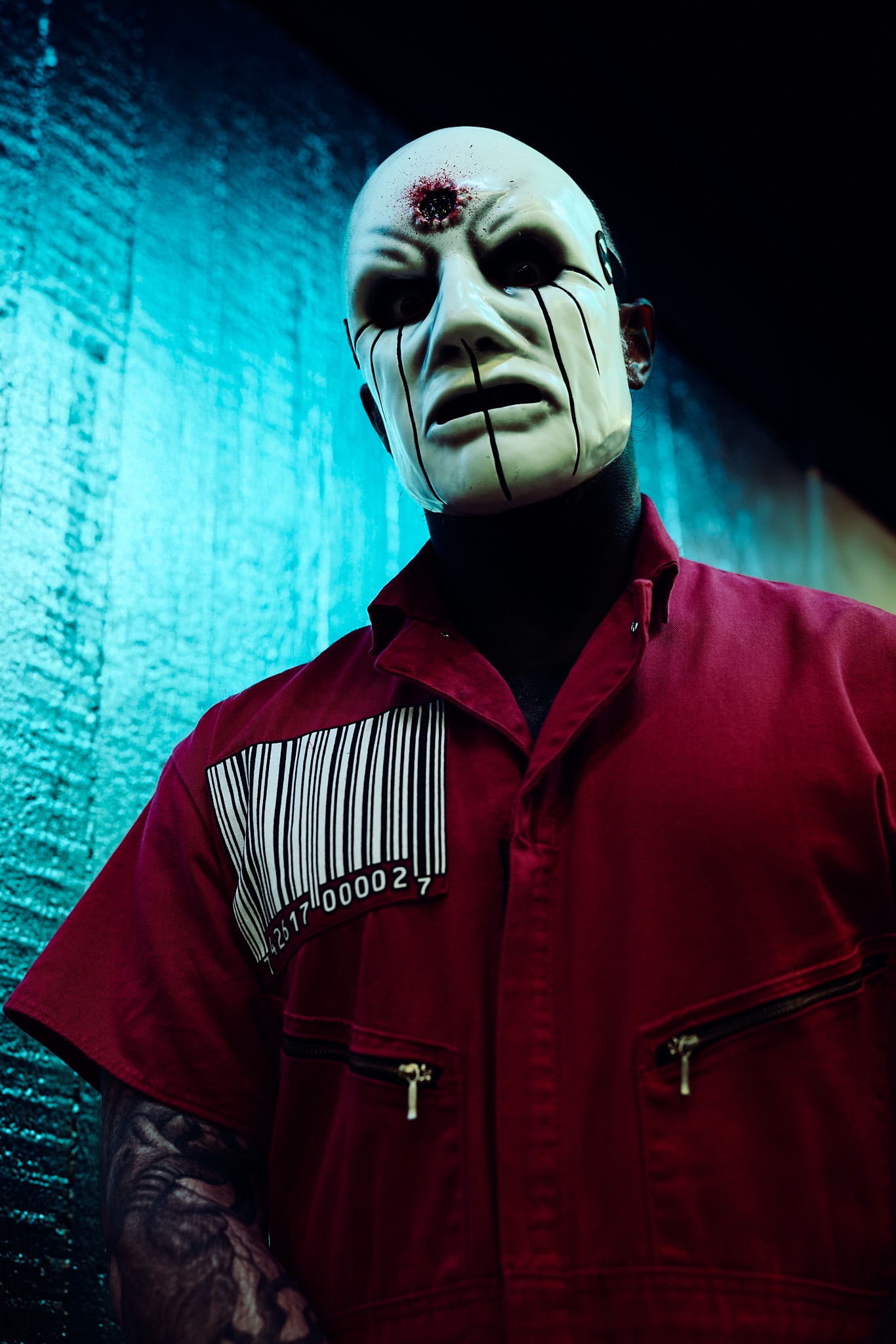 Une personne portant un masque blanc de style Slipknot avec des lignes noires, une combinaison rouge avec un grand code-barres, se tient contre un mur texturé bleu et vert.