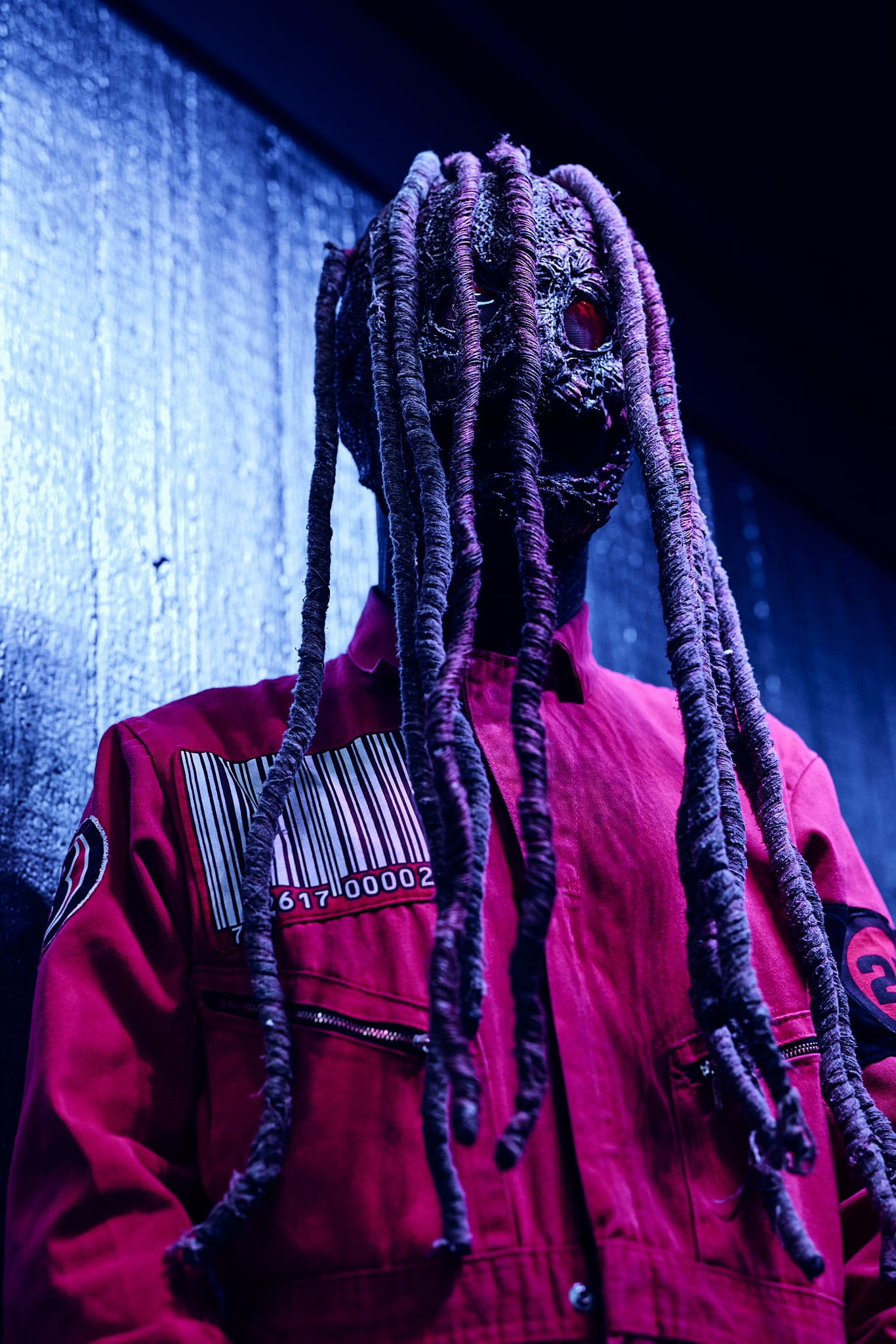 Une personne vêtue d'une combinaison rouge avec un code-barres et des patchs se tient contre un mur sombre, le visage caché par un masque inspiré de Slipknot avec de longues extensions ressemblant à des dreadlocks et un seul œil rouge incandescent.