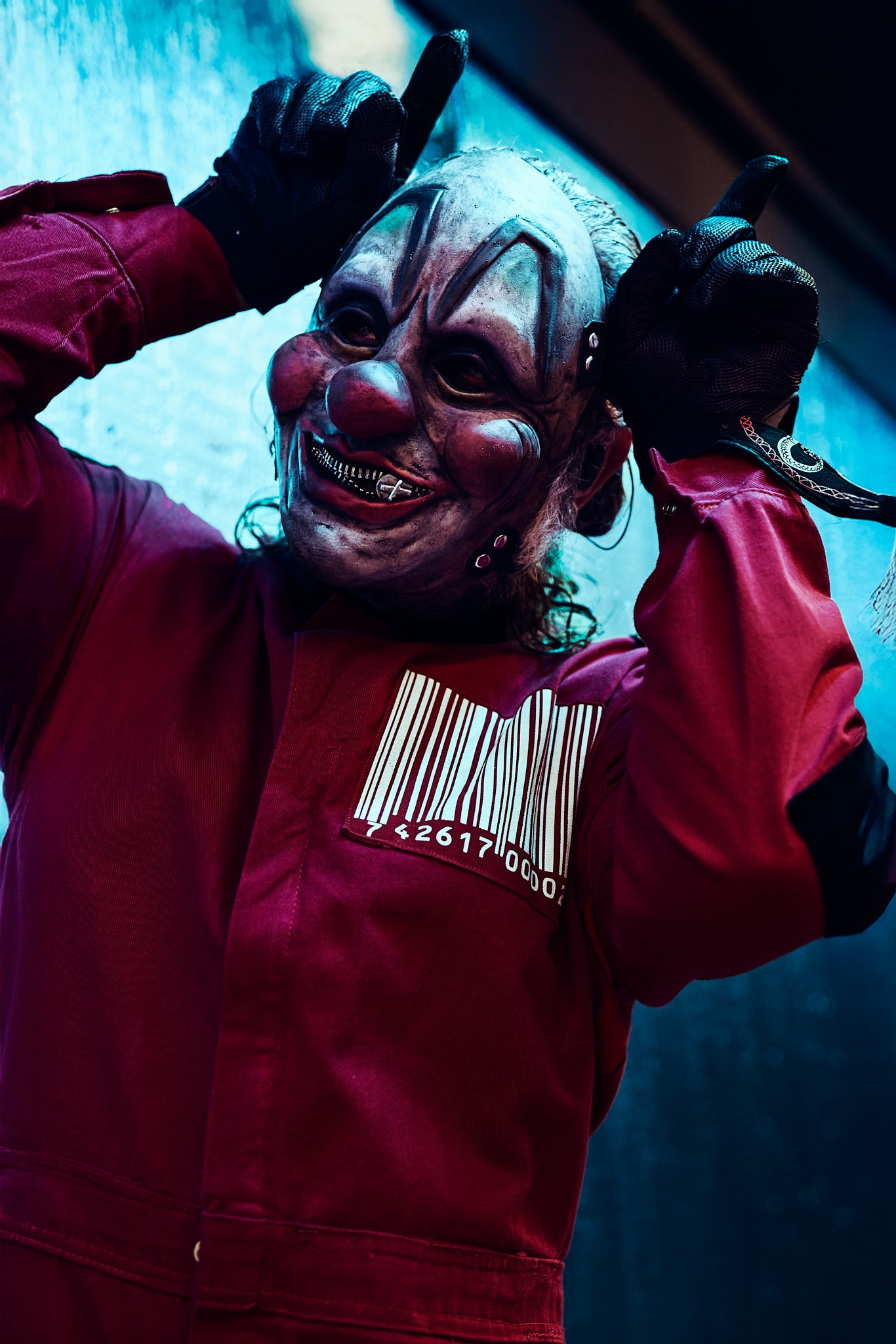 Une personne portant un masque de clown sinistre et une combinaison rouge avec un code-barres, rappelant le style emblématique de Slipknot, tient ses mains gantées près de sa tête, imitant des cornes, sur un fond sombre et lunatique.