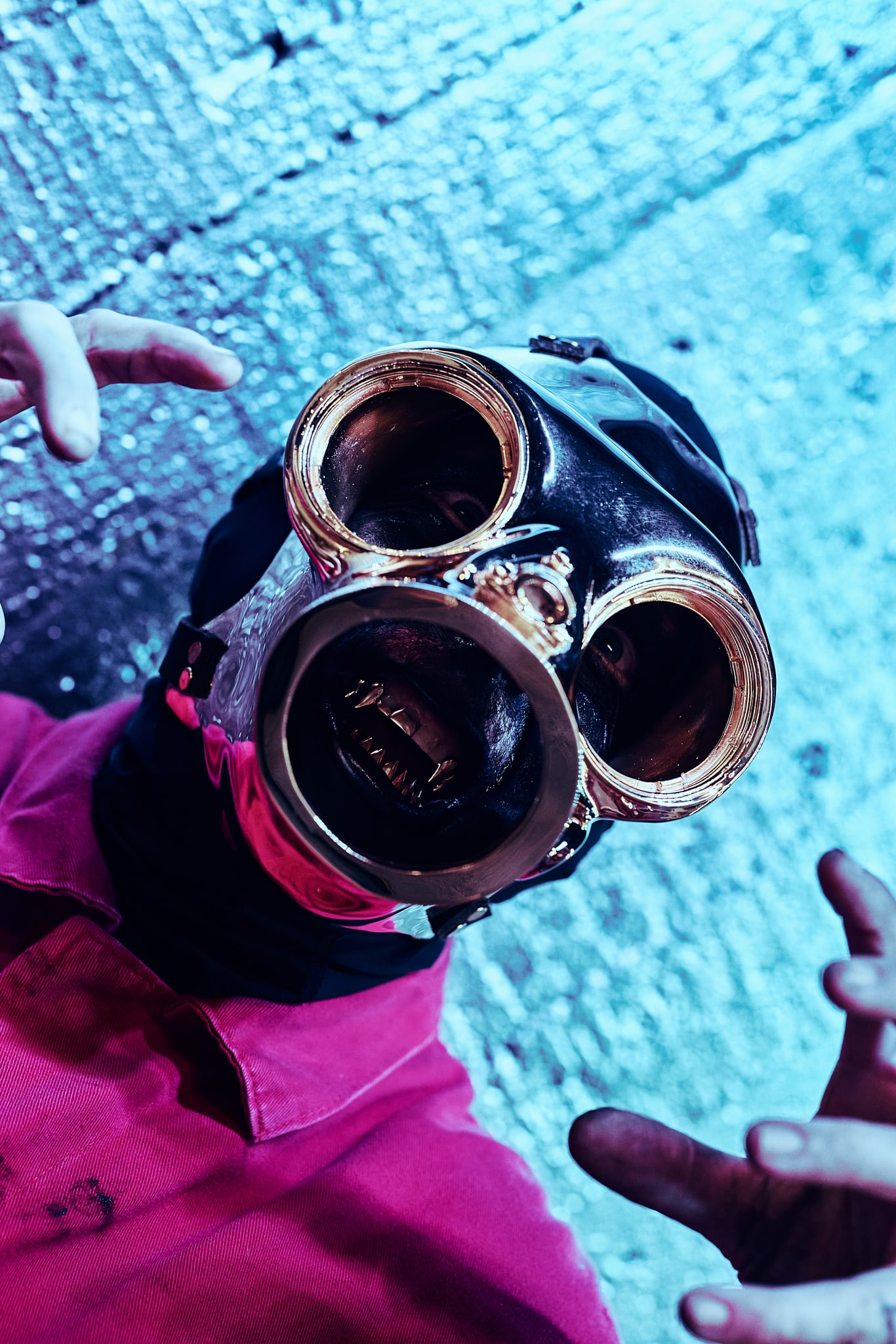 Une personne portant un masque à gaz métallique brillant avec de grandes lentilles rondes et une chemise rouge, rappelant le style énervé de Slipknot, fait des gestes avec ses mains. L'arrière-plan est texturé et éclairé par une lumière bleue.