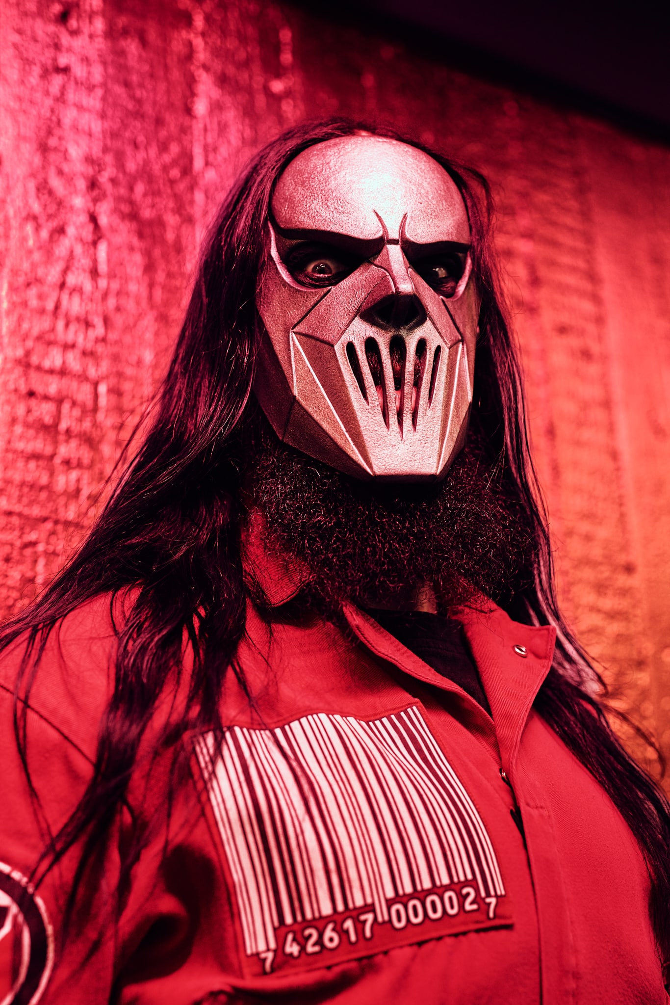 Une personne aux longs cheveux noirs et à la barbe fournie porte un masque métallique à angles vifs inspiré de Slipknot et une combinaison à code-barres, sur un fond texturé et éclairé en rouge.