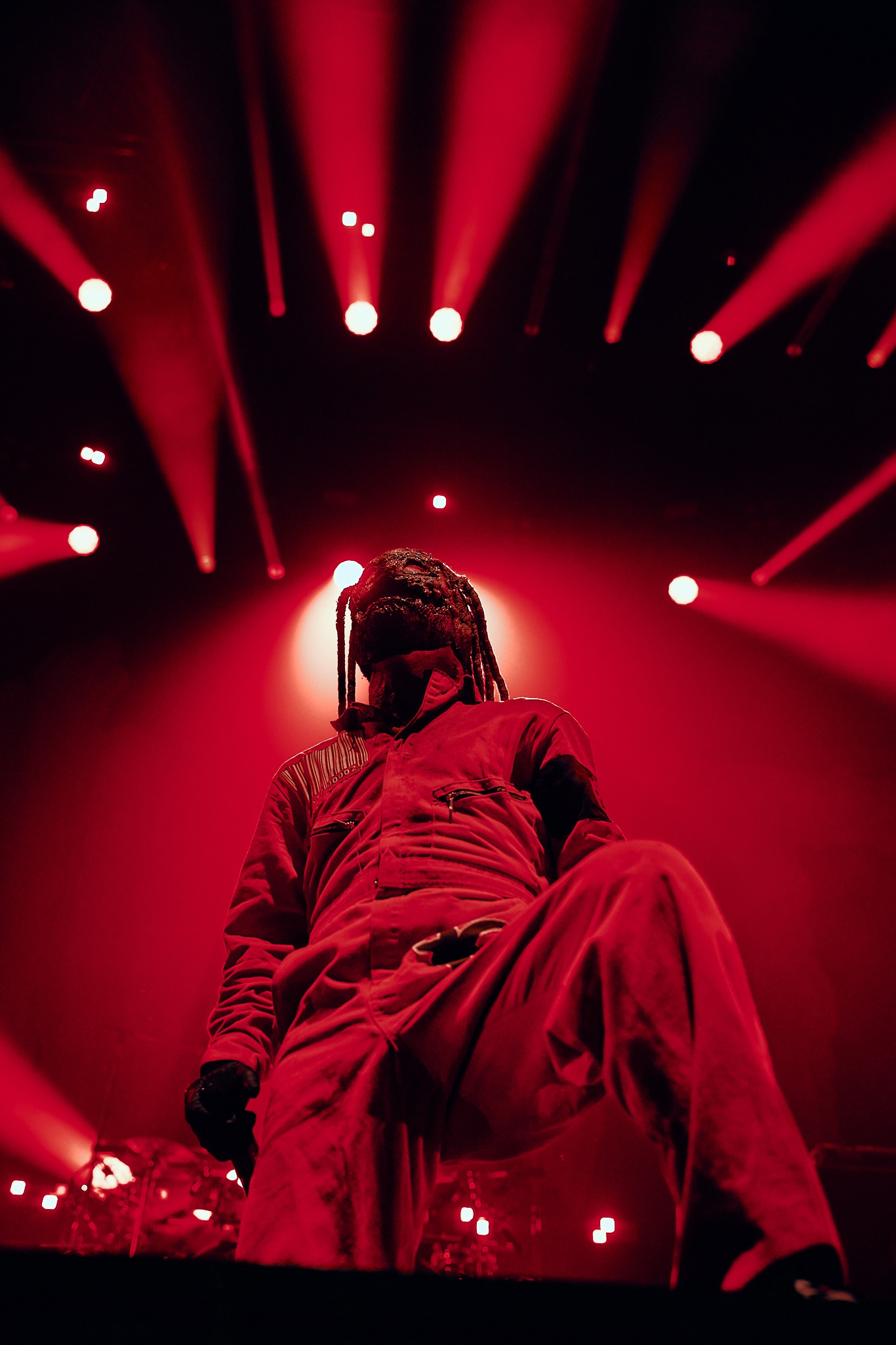 A person wearing a red jumpsuit and mask stands on stage under dramatic red lighting, with spotlights shining above and behind them, evoking the intense atmosphere of a Slipknot concert.