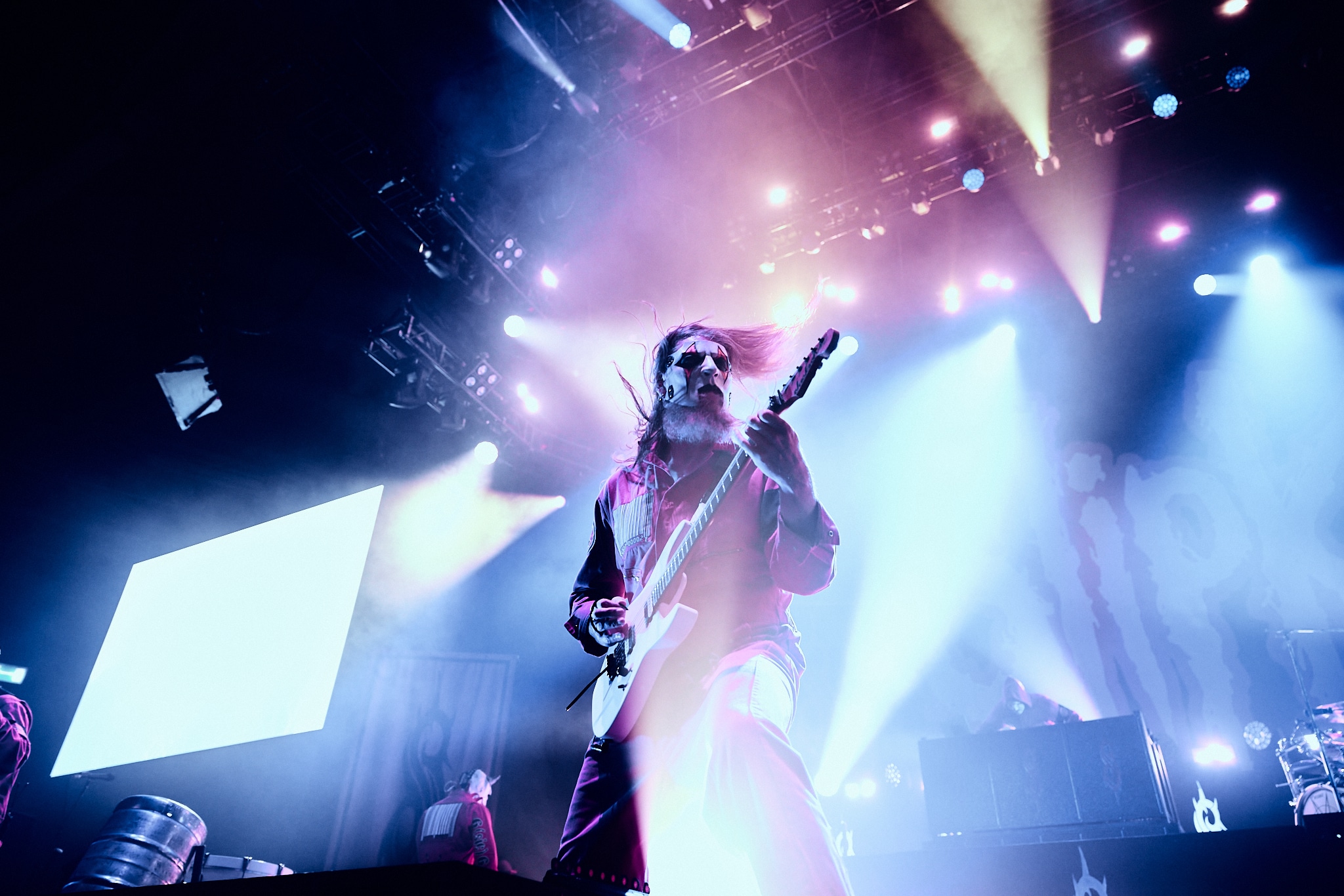 Un guitariste vêtu d'un costume et d'un masque joue énergiquement sur une scène très éclairée, entourée de lumières et d'effets colorés, avec le nom de Slipknot partiellement visible en arrière-plan.