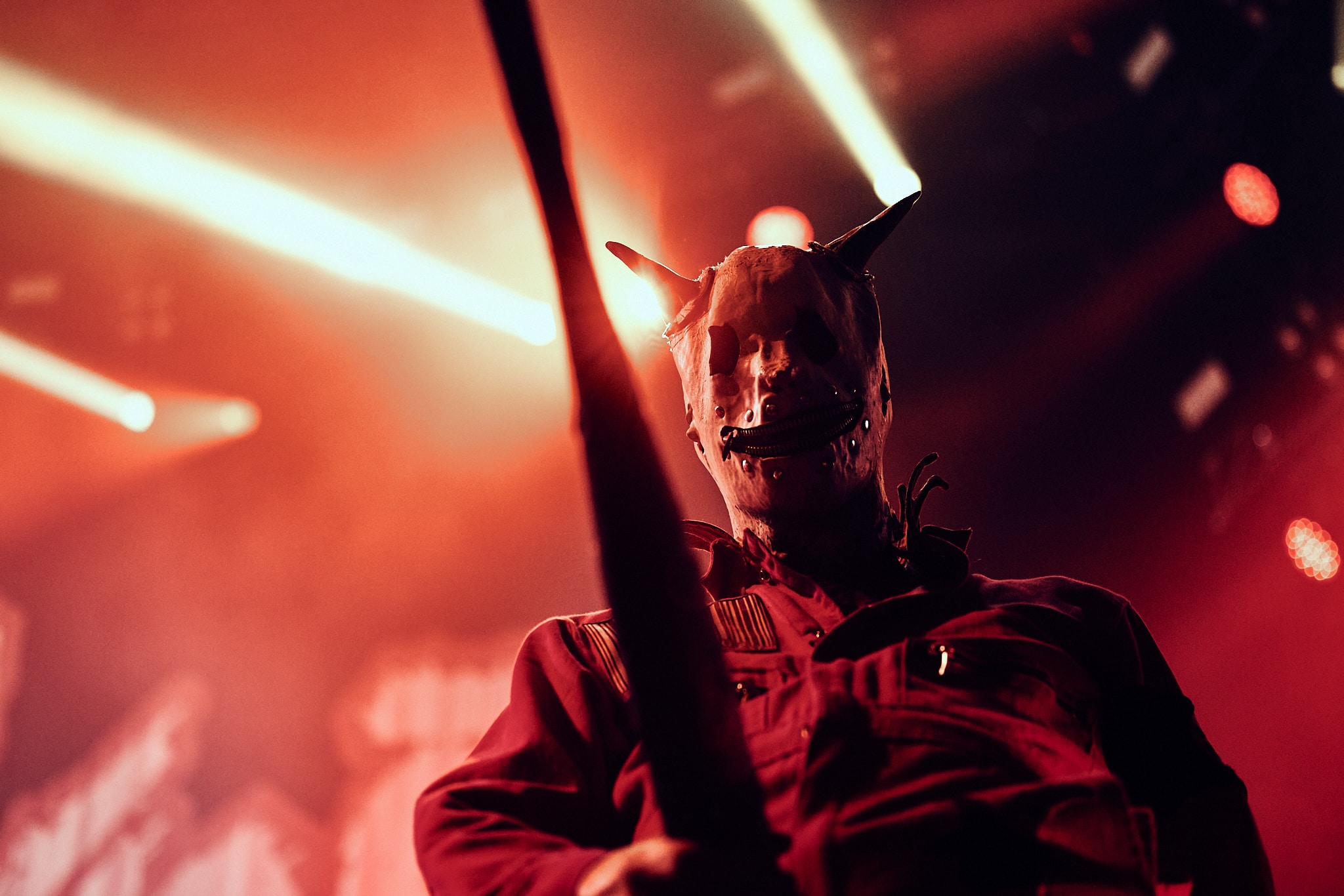 Un artiste portant un masque à cornes et une tenue rouge se tient sur scène sous un éclairage dramatique, tenant un objet ressemblant à une chauve-souris - évoquant l'énergie intense de Slipknot - tandis que des projecteurs lumineux traversent l'atmosphère enfumée de l'arrière-plan.