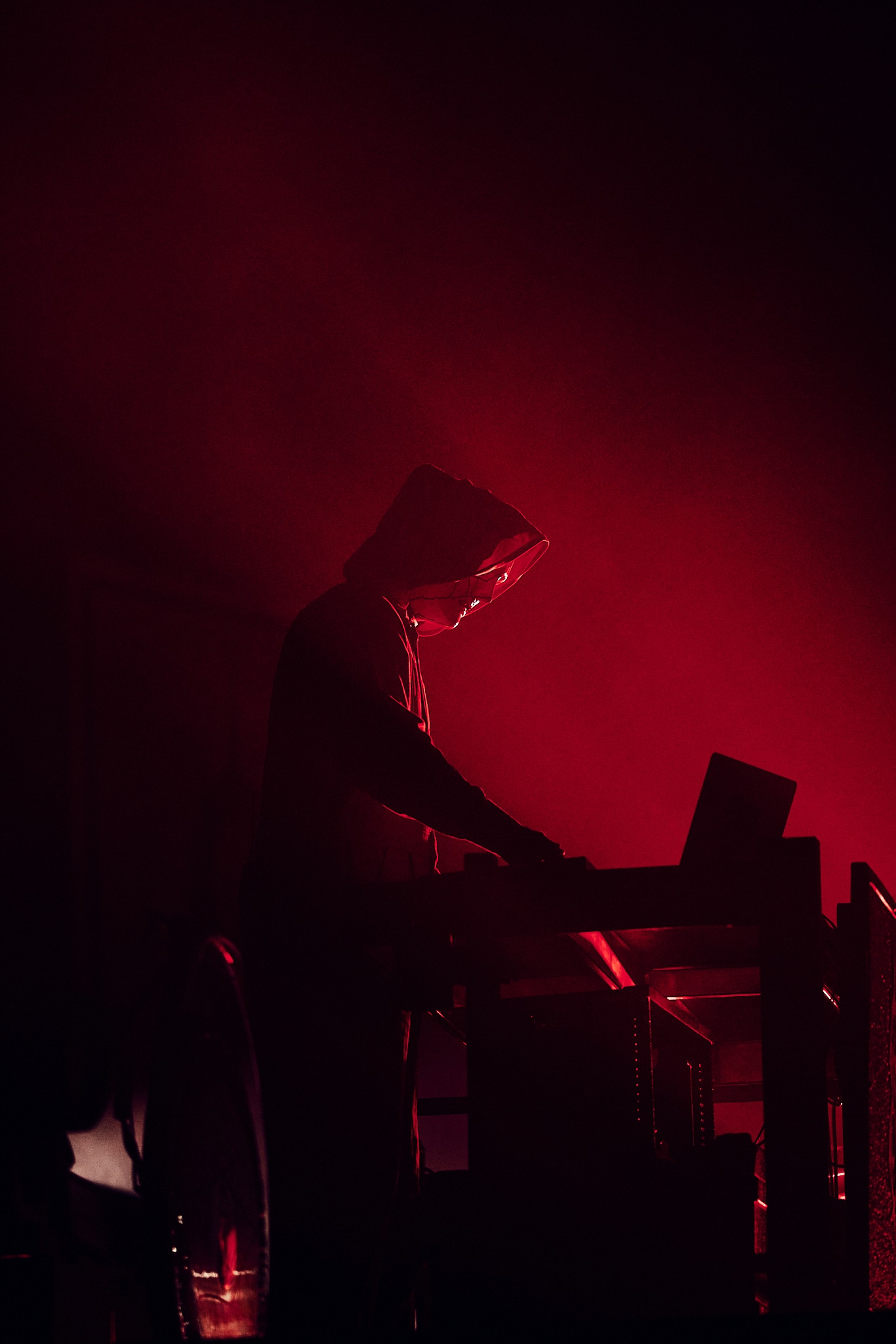 Une personne portant une cagoule se tient devant un ordinateur portable, éclairé par des lumières rouges dramatiques et de la fumée sur une scène sombre, évoquant l'atmosphère intense et mystérieuse d'un concert de Slipknot.