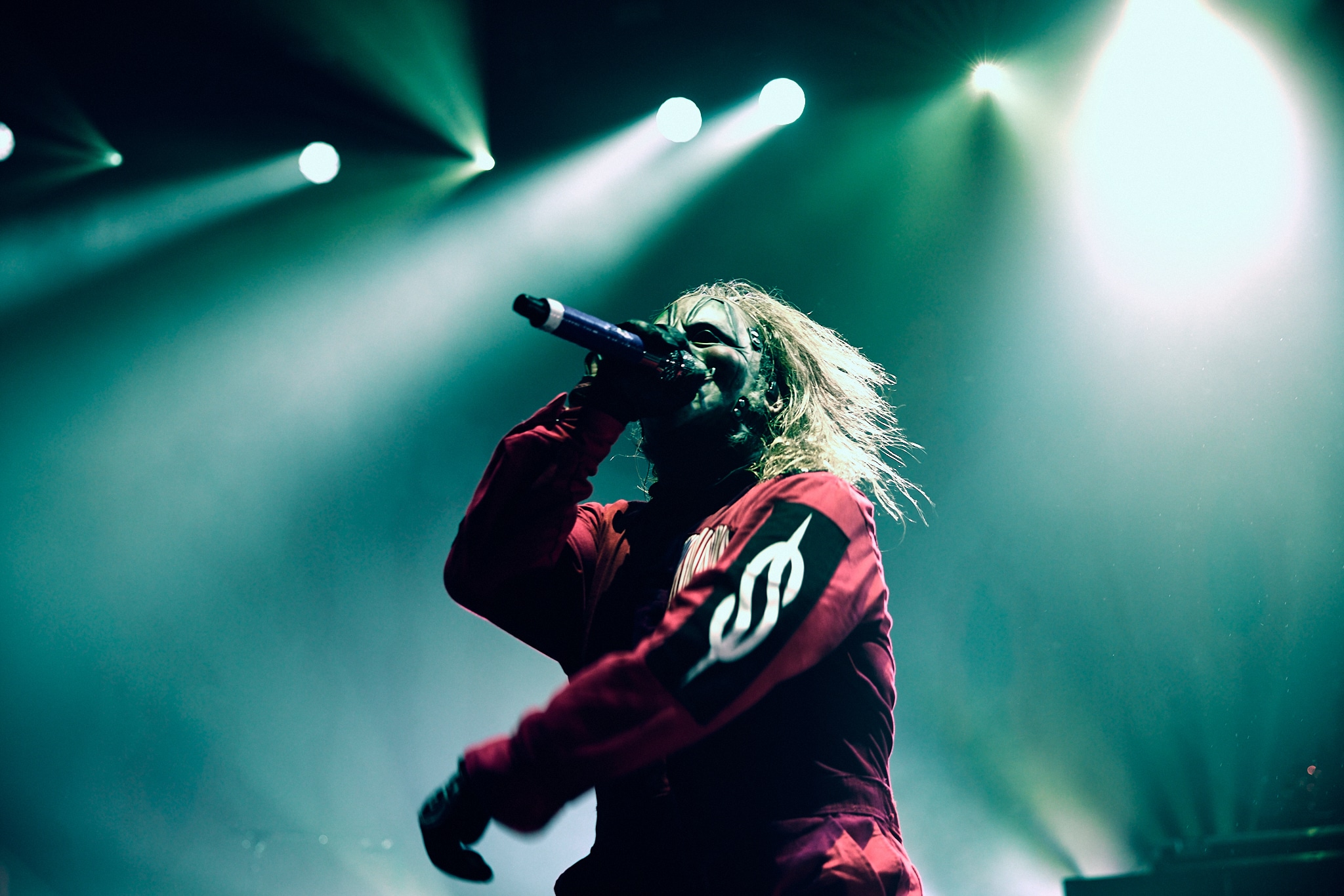 Un artiste vêtu d'une veste rouge avec un logo blanc chante avec passion dans un micro sur la scène, éclairée de façon spectaculaire par des lumières colorées et entourée de fumée, évoquant l'intensité de l'énergie d'un concert de Slipknot.